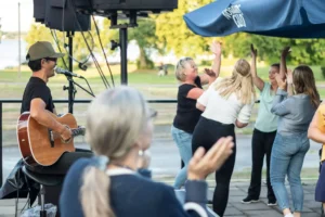 Musiker spelar gitarr och sjunger medan publiken dansar och applåderar under ett utomhusevenemang vid vattnet.